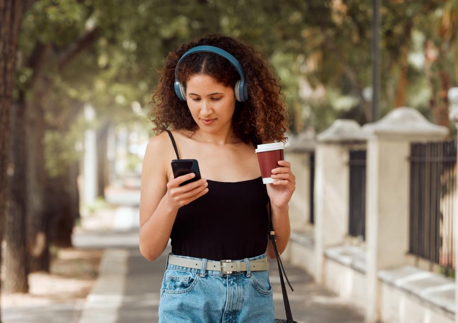 A young woman walking down the street in headphones, reading from her phone