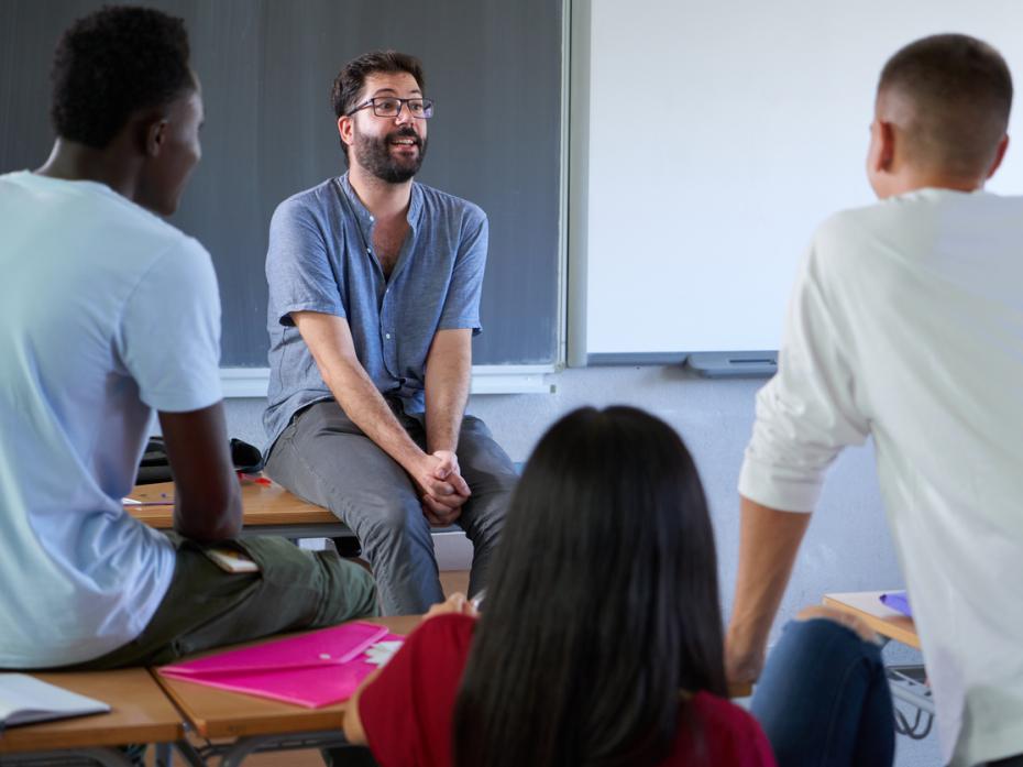 A lecturer sitting on a desk speaking to students in an informal manner