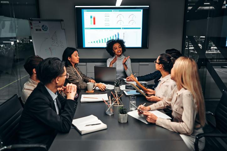 In a boardroom, a group of women discuss the charts on the screen