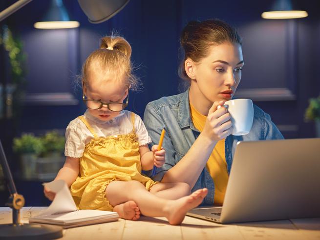 A woman sits at a laptop with a small toddler on the desk next to her