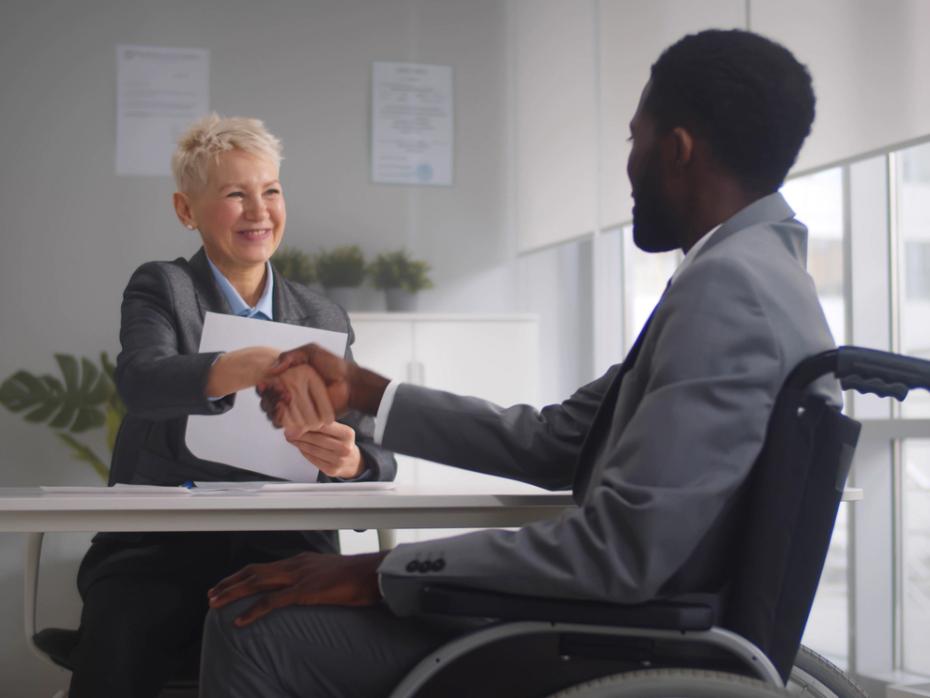 Black male job candidate in a wheelchair shaking hands with mature white woman