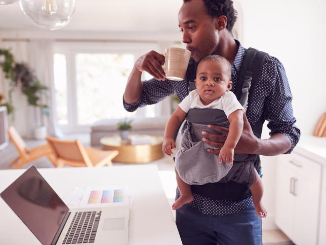 A man with a baby in a sling checks his laptop while drinking coffee