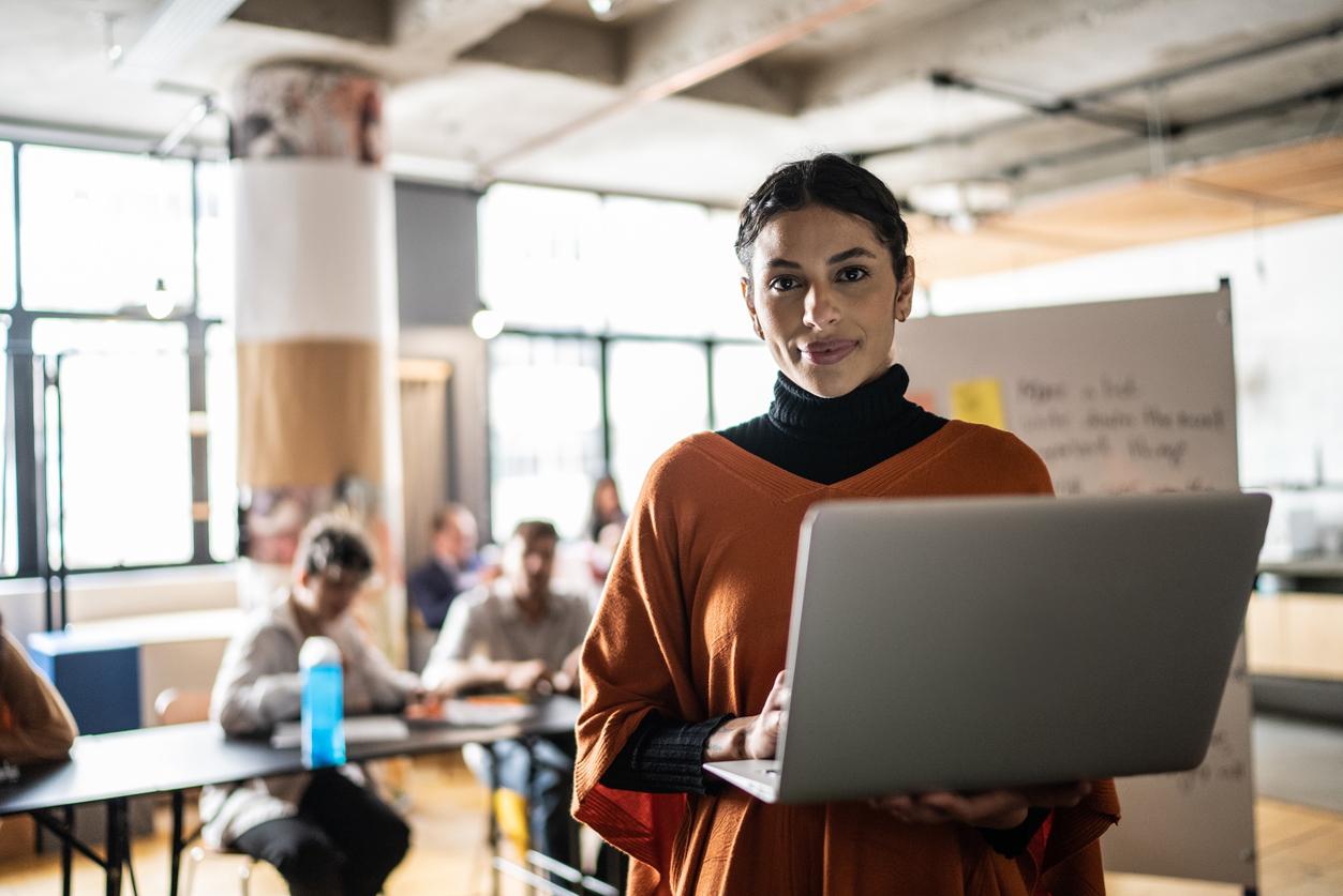 A teacher in her 20s holding a laptop smiling at the camera while her students work at desks behind her