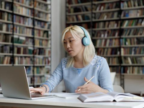Young Asian woman working at laptop in library wearing headphones