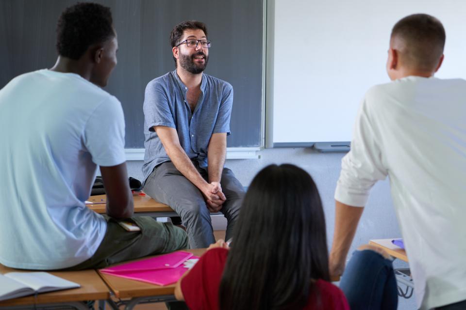 A lecturer sitting on a desk speaking to students in an informal manner