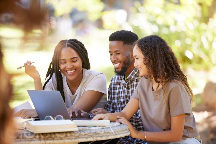 A group of students sit happily around a laptop in the sunshine