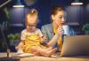 A woman sits at a laptop with a small toddler on the desk next to her